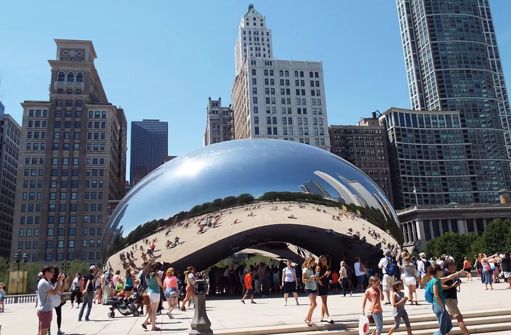 A photograph of the Cloud Gate sculpture in Chicago on a sunny day. The cloud Gate is a huge polished metallic coffee bean that reflect the sky scrapers in ithe back ground