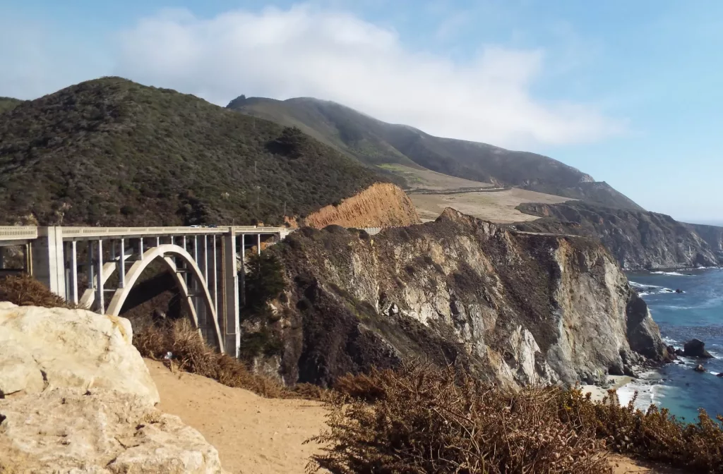 A photgraph of the Bixby Bridge on the Big Sur on Highway one in California with mountains and cliffs to teh rear and the the pacific ocean in the foreground