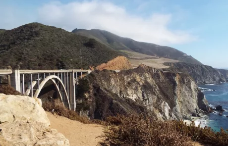 A photgraph of the Bixby Bridge on the Big Sur on Highway one in California with mountains and cliffs to teh rear and the the pacific ocean in the foreground